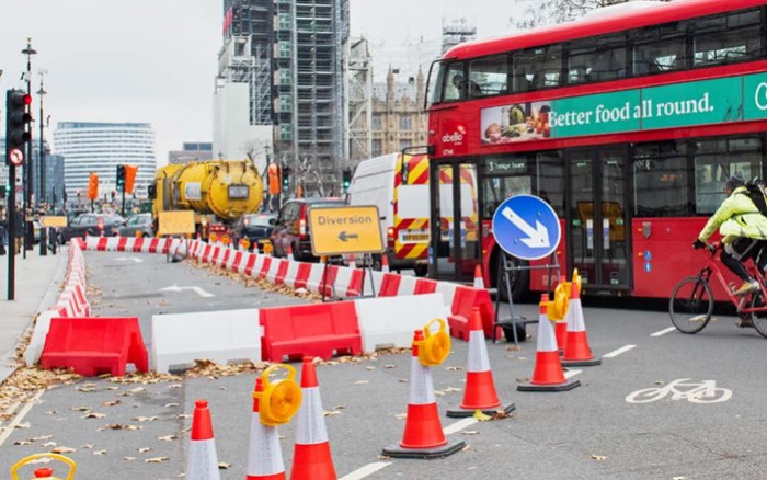 Road works site with traffic management placed on the left side of the carriageway, removing a lane and directing traffic to the right of the works, along with a diversion sign. Cars, a bus and a cyclist visible in image